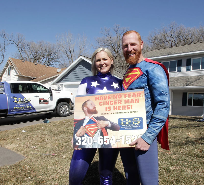 Emily & Jeremy Salzbrun dressed in superhero costumes advertising the H&S Heating & Air Conditioning business in front of a residential home.